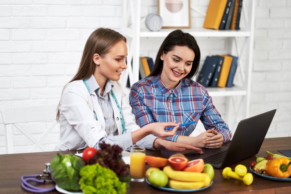 Two women at a table with a laptop, exploring weight loss options, accompanied by a selection of fruits and vegetables.
